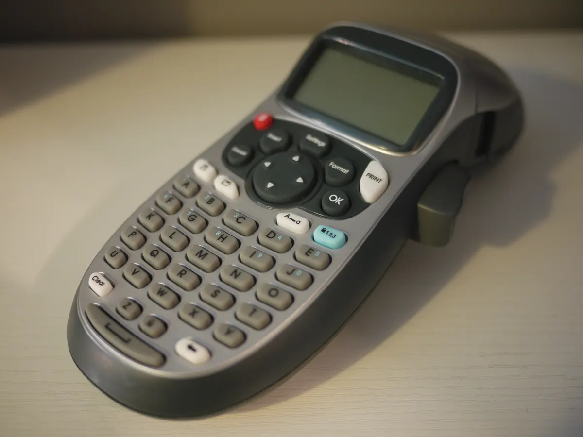 A gray DYMO LetraTag 100H handheld label maker with an alphabetical ABC keypad and a small LCD screen on a white desk